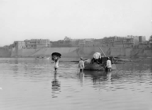 boat and men wading in water with buildings on the bank