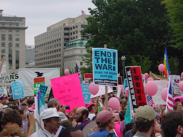 A group of protestors march together carrying signs about ending a war and bringing the troops home.