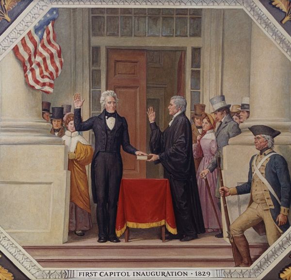A detailed painting captures the outdoor inauguration of Andrew Jackson as president. He stands prominently in the center, dressed in a dark suit, with his right hand raised in oath while facing a robed official holding a book.