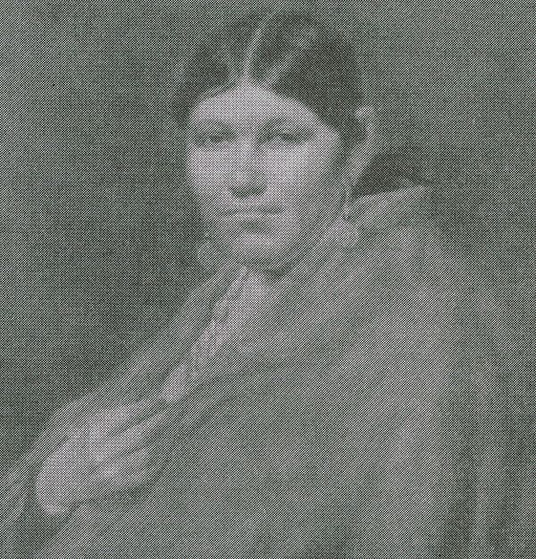 A grainy, black-and-white portrait shows a young Cherokee woman gazing directly at the viewer. She is draped in a blanket or shawl and wears a necklace with what appears to be shells or beads.