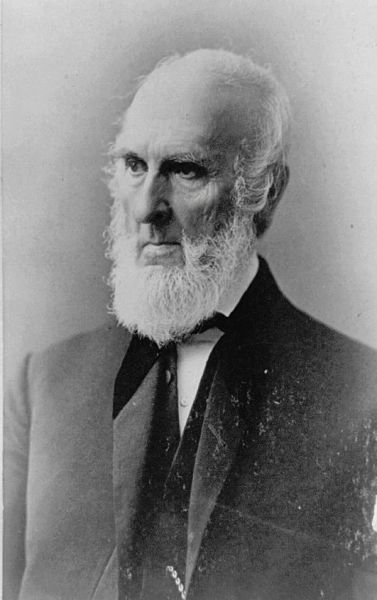 A black and white photograph of John Greenleaf Whittier, a 19th-century American Quaker poet and abolitionist. He is depicted in a formal suit and bow tie, with a receding hairline and a full, long white beard, looking off to the left with a serious expression.