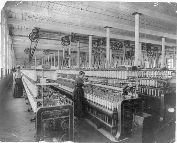 A vintage, black-and-white photograph shows the long, wide interior of a textile mill, filled with rows of large spinning machinery and overhead power belts and pulleys. Two female workers in long dresses attend to the machines, with one in the foreground tending to a bank of spindles loaded with yarn.