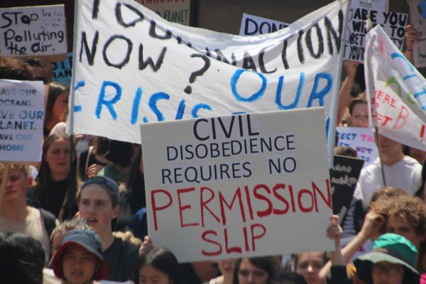 A photograph showing a crowd of diverse young people participating in a climate change protest. The foreground features a sign reading, "CIVIL DISOBEDIENCE REQUIRES NO PERMISSION SLIP," with other signs and banners visible in the background.