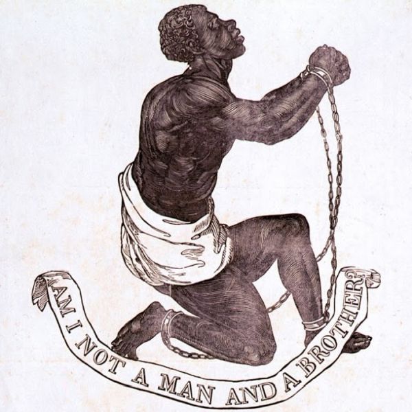 A black and white 18th-century abolitionist medallion design depicting an enslaved Black man kneeling on one knee, chained at the wrists and ankles, looking up. A scroll beneath him reads the famous abolitionist motto: "AM I NOT A MAN AND A BROTHER."