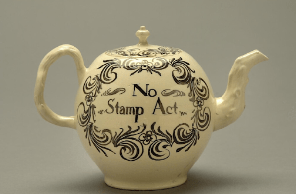 A white ceramic teapot is decorated with black floral and scroll patterns. The center of the teapot features a prominent decorative cartouche with the text, "No Stamp Act."