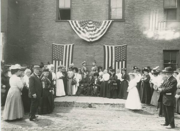A black and white historical photograph shows a large group of people, primarily women in late 19th or early 20th-century attire, standing on steps in front of a brick building. The building is decorated with two vertically hung American flags and a pleated bunting across the top, indicating a patriotic gathering or event.