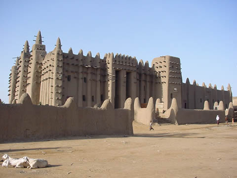 Photograph of imposing building surrounded by a mud wall, with goats in the foreground