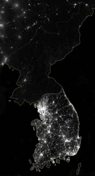 a satellite image of the Korean Peninsula at night, showing heavy lighting in the south and vast darkness in the north