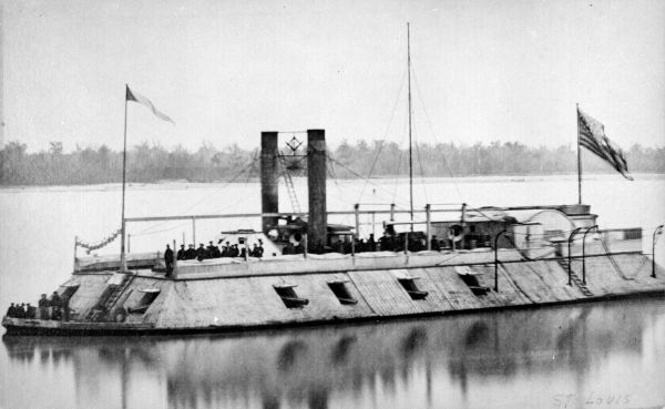 In this black-and-white photograph, a Union ironclad warship sits in calm water with an American flag flying from its stern and a smaller flag at the bow. The ship features a flat, sloped armored deck with several open gun ports along its side and two tall, dark smokestacks rising from the center.