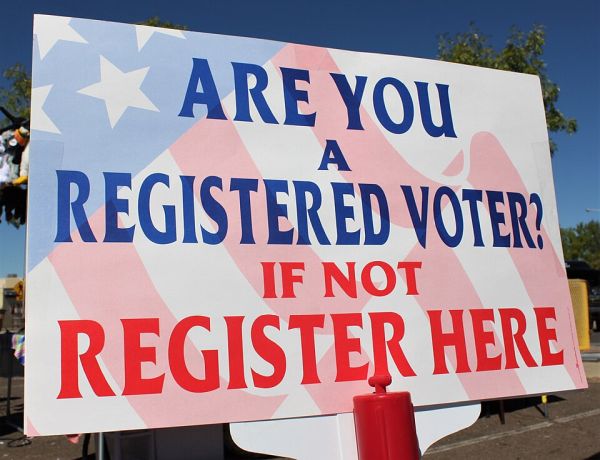 A bright, close-up photograph shows a large white sign with red and blue lettering that asks, "Are you a registered voter? If not, register here." The background of the sign is a stylized American flag with stars on the top left and red and white stripes covering the rest.