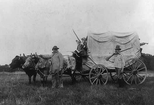 photograph of a covered wagon pulled by oxen; two women sitting on wagon, two men walking alongside it