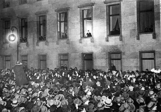 photograph of Hitler standing in a lit window of a building, crowds below in the street