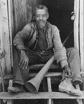 old photograph of a Black man sitting on a wooden step, holding a large horn
