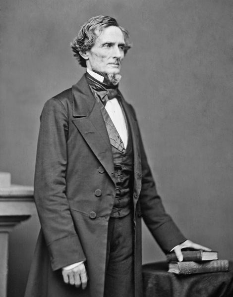 This black-and-white full-length portrait shows Jefferson Davis standing and looking toward the right with a serious expression. He wears a dark frock coat over a matching vest and white shirt, with one hand resting on a pair of books on a small table.