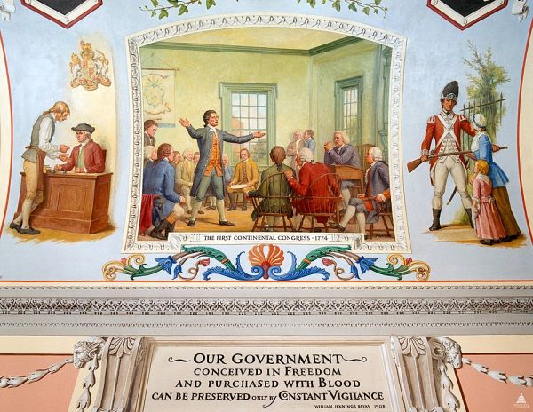 An oil painting shows a man in a blue coat standing and passionately speaking to a group of seated men in a wooden room. Most of the men in the painting are wearing jackets, breeches, and white shirts with ruffled cuffs.