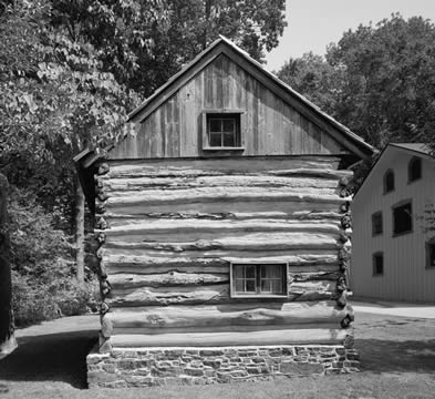 black and white photo of a colonial log cabin, one end with two windows but no doors