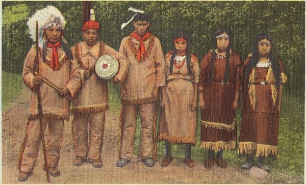A faded, postcard-like photograph shows six Indigenous people standing in a line outdoors. They are dressed in traditional clothing, with some wearing headdresses and holding accessories like a ceremonial staff and a drum.