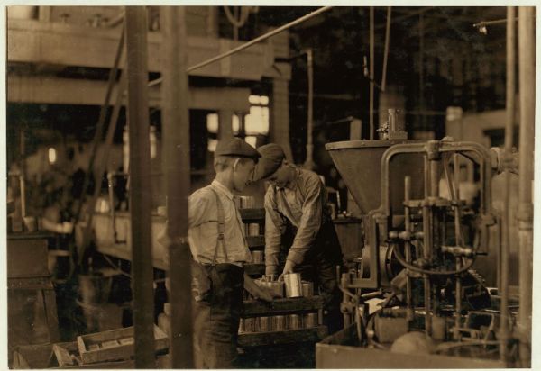 A historical black-and-white photograph shows the dimly lit, machinery-filled interior of a canning factory, where two young male workers wearing caps and work clothes are busy near an industrial machine. One boy in suspenders is holding an empty can while the other is leaning over a rack of cans being processed by the large piece of equipment.