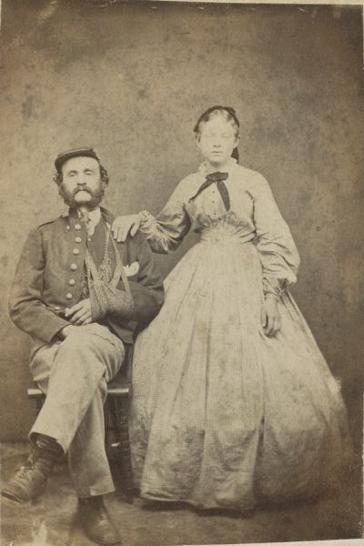 In this sepia-toned studio portrait, a Confederate soldier with a thick beard and mustache sits with his legs crossed, his right arm resting in a patterned sling. His wife stands beside him with her hand on his shoulder, wearing a long-sleeved, high-collared dress with a full, checkered skirt and a small dark bow at her neck.