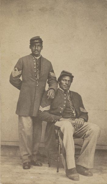 A sepia-toned photograph shows two Black men in Union Army uniforms, one standing and one seated. Both men wear caps and coats with sergeant's chevrons on the sleeves, posing together for a formal portrait.