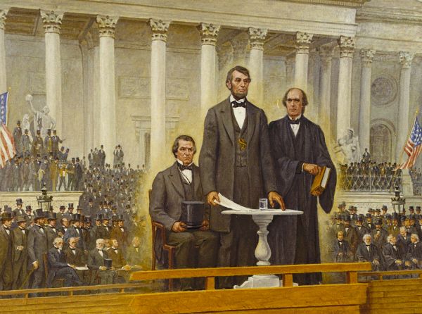 Photograph of Abraham Lincoln standing on a raised platform at the U.S. Capitol during his second inauguration in 1865, surrounded by a large crowd of officials and spectators. Lincoln faces forward as he delivers his speech, with columns and architectural details of the Capitol visible behind him.