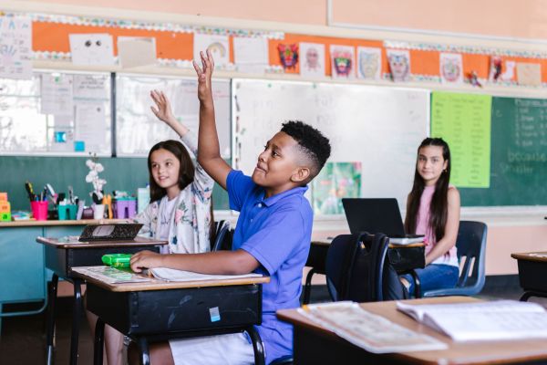 In a brightly lit classroom, a young Black boy in a purple polo shirt sitting at his desk eagerly raises his hand to answer a question.