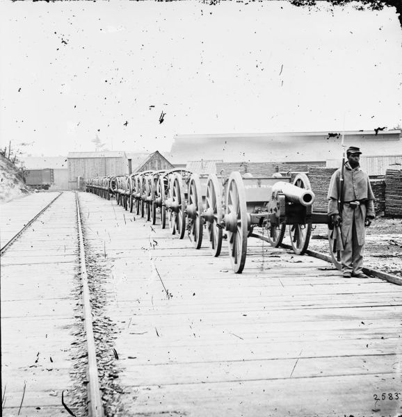 In this black-and-white photograph, a long line of cannons on large wooden wheels stretches into the distance along a wooden platform next to a set of railroad tracks. A Black soldier in a Union military uniform and overcoat stands at attention on the right, holding a rifle with a fixed bayonet next to the lead cannon.