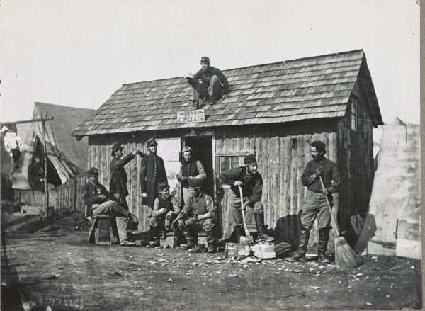 In this black-and-white photograph, a group of Union soldiers in various uniforms and caps is gathered around a small, rustic wooden cabin labeled "Pine Cottage." One soldier sits perched on the shingled roof while others stand or sit below, with two men in the foreground engaged in chores like sweeping and chopping wood.