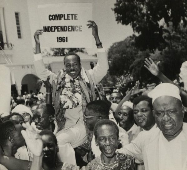 Black-and-white photograph of a crowd celebrating independence. A smiling man raised above the crowd holds a sign reading “Complete Independence 1961,” while people around him cheer and raise their hands.
