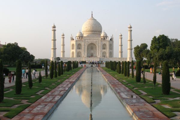 Front view of the Taj Mahal, a white marble mausoleum with a large central dome and four tall minarets, reflected in a long rectangular pool surrounded by formal gardens and visitors.