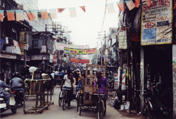 Busy urban street in India lined with shops and signs, with pedestrians, bicycles, rickshaws, and motorcycles moving through a crowded commercial area decorated with strings of colorful flags.