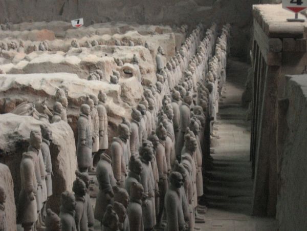 Rows of life-size Terracotta Army soldiers stand in excavated pits, with numerous clay figures arranged in formation beside earthen walls and walkways.